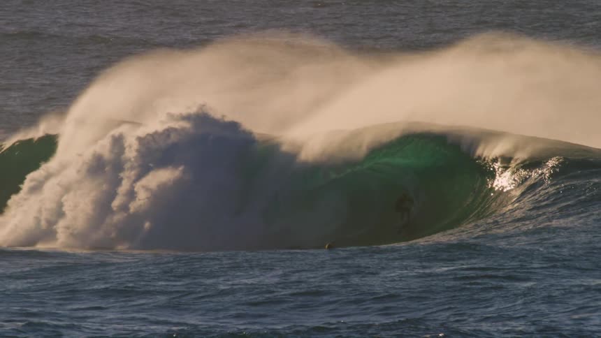 A surfer is riding a large powerful wave with a dramatic crest of white foam with other surfers visible in the background