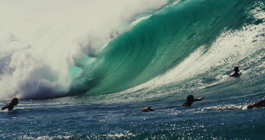 A big wave is breaking near some surfers who are paddling their boards forward