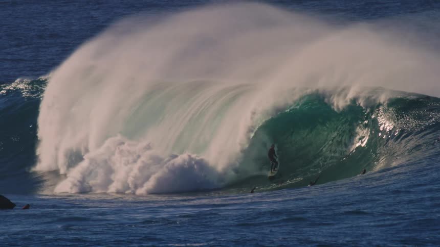 A large waves rolls over as a man rides it and people look on