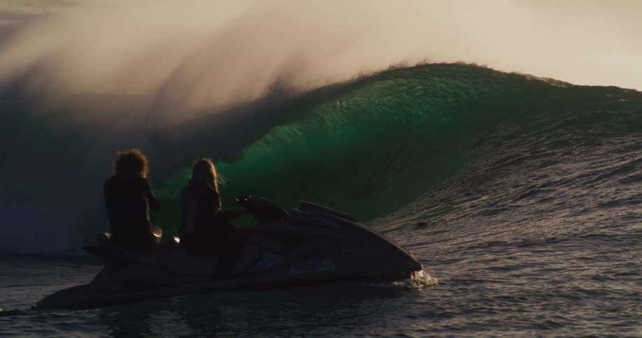 A big ocean wave breaking at its crest and there are some people on jet skis next to it