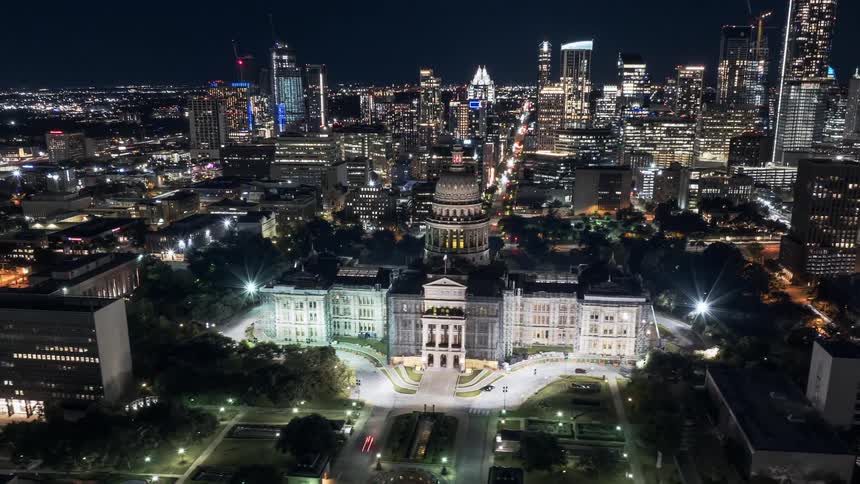 Aerial night view of a large illuminated neoclassical building surrounded by gardens with a city skyline in the background