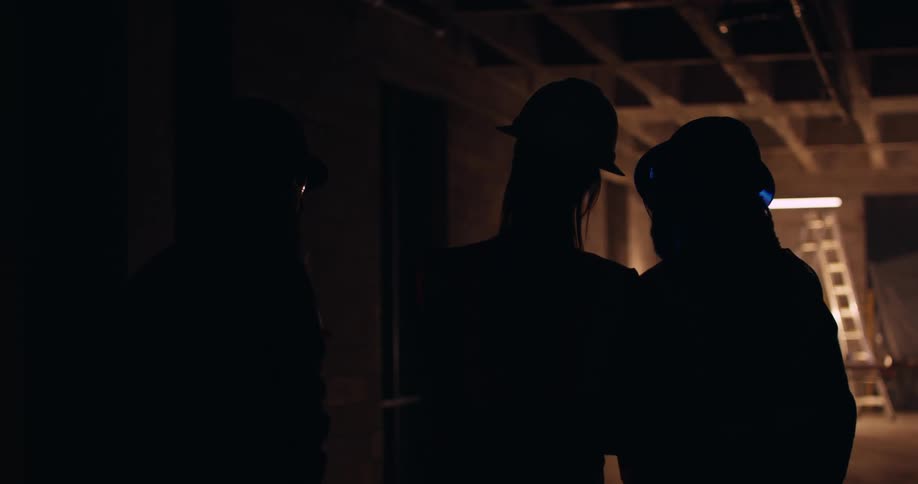 The silhouettes of three people standing in a dark construction area wearing hard hats and talking as they point out details in a building site.