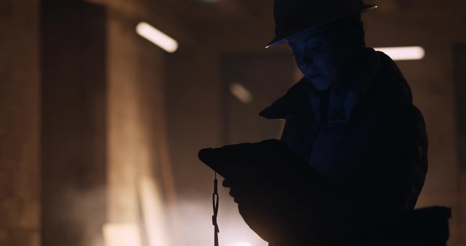 Silhouette of a female engineer in a hard hat and safety gear holding and looking down at a tablet while standing in a dusty and dimly lit construction site at night.
