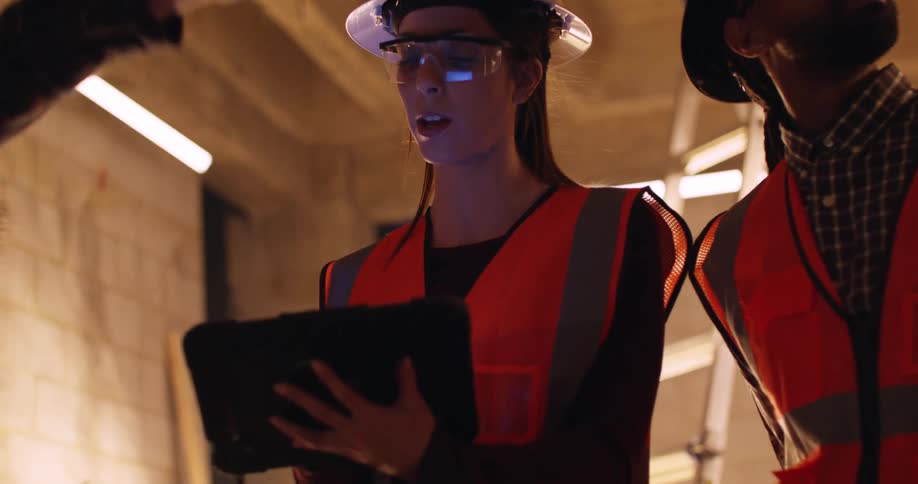 A female engineer is using a tablet to inspect a construction site along with two other male colleagues, all wearing safety gear and hard hats.