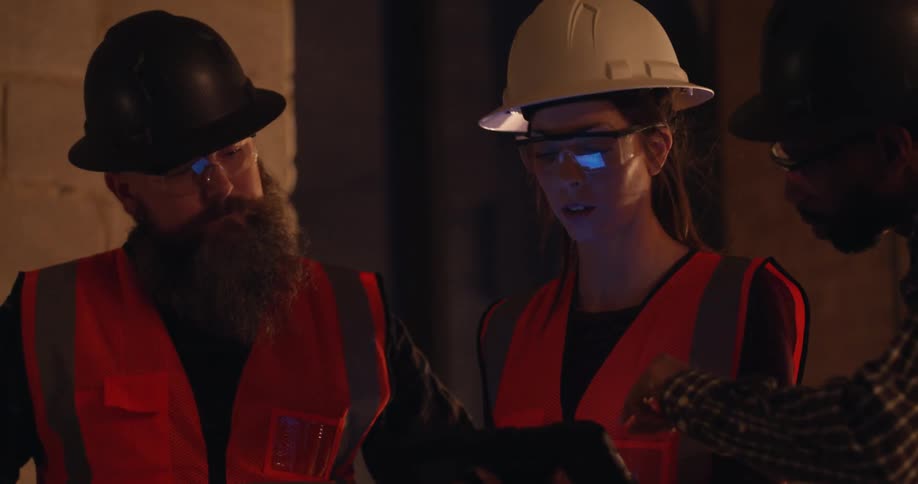 Three engineers in hard hats and safety gear are having a discussion over project plans on a tablet while standing in a building under construction at night.