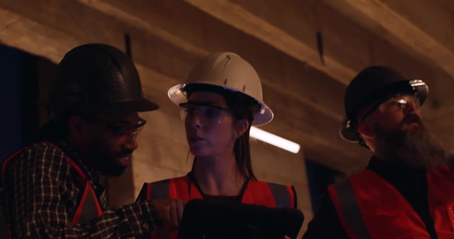 Three engineers wearing reflective vests and safety equipment analyze and discuss the structure of a construction site while consulting the plans on a tablet.