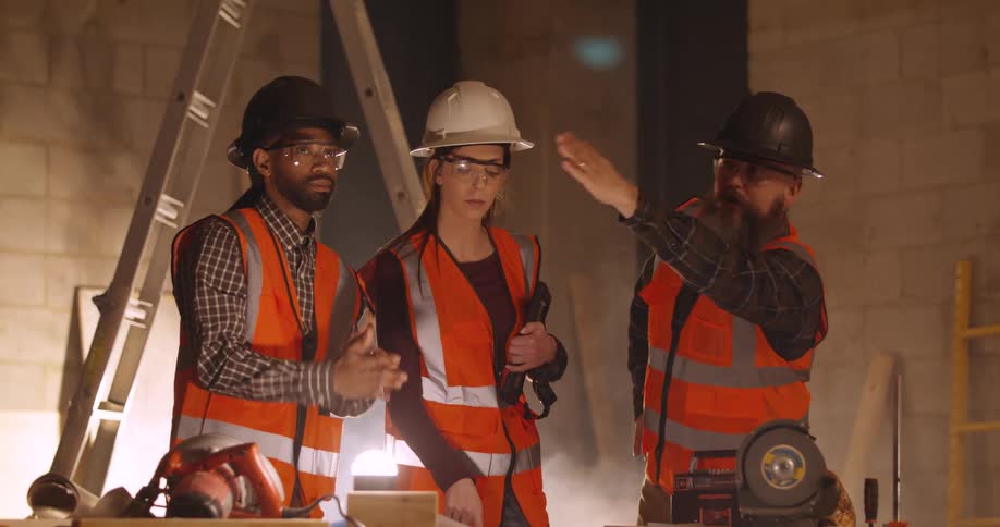 On a construction job site three workers including a bearded white man a woman and an african american man are in the midst of planning and discussing their next steps as the bearded man gestures with his hand to signal the teams collaborative appro