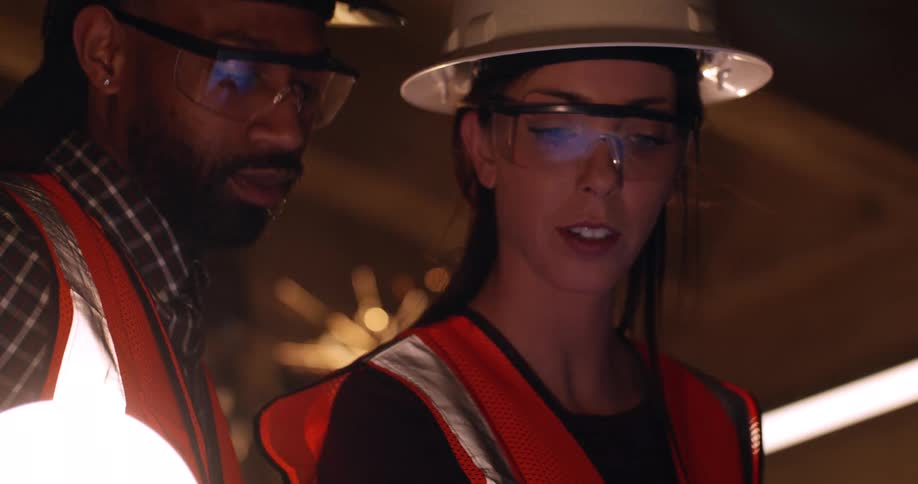 Close up of a woman and a black man wearing hard hats and safety gear as they discuss building project plans while standing in an active construction site with a welder producing sparks behind them.