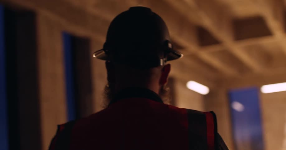 A man with a beard wearing safety gear and a hard hat is walking through an indoor construction site at night and pointing out different spots in progress.