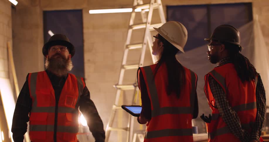Three diverse civil engineers walk through and inspect progress at a construction site at night, with a floodlamp as the main source of light.