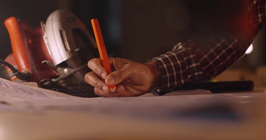 The hand of a black man holding a square pencil and writing notes on a set of project plans on a table with a circular saw and other tools scattered around.