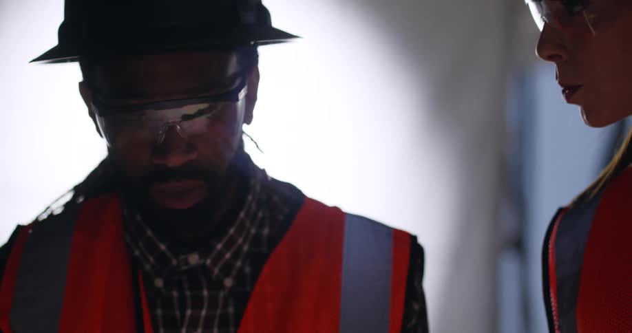 A black male civil engineer is looking down and nodding while discussing a building project with a female worker standing next to him in a construction site, with both of them wearing safety gear for protection.