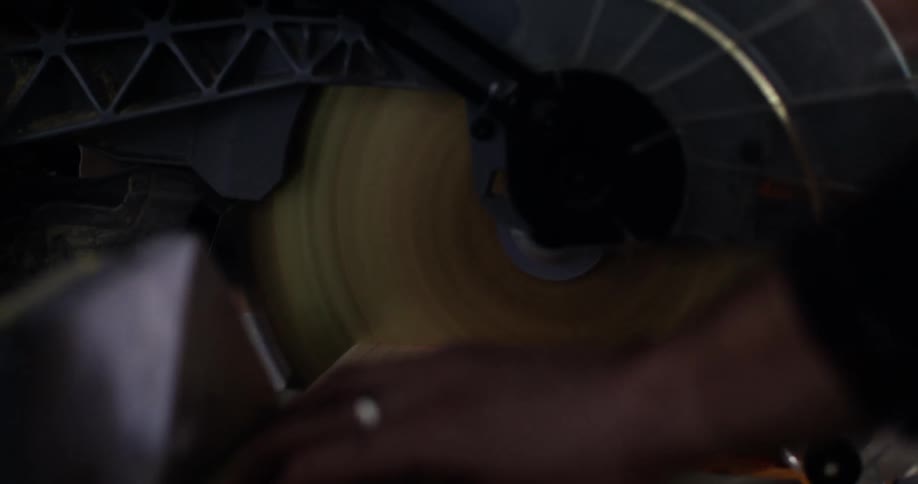 Close up of a man's hand holding a piece of wood and using the rotating blade of a miter saw to cut the wood to size in his workshop