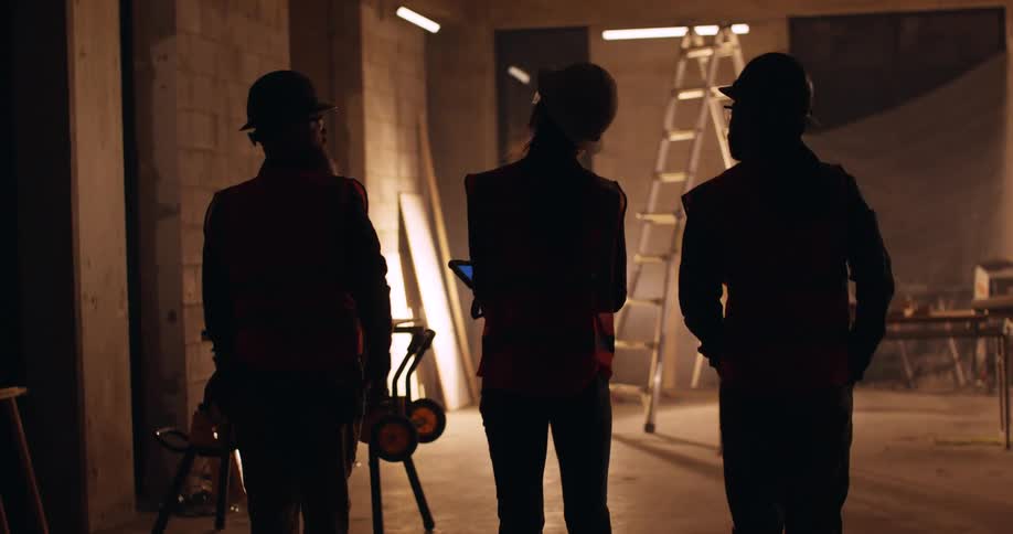 View from behind of three civil engineers, two men and a woman, discussing a construction project and walking the site at night, illuminated only by a floodlight and a few lanterns.