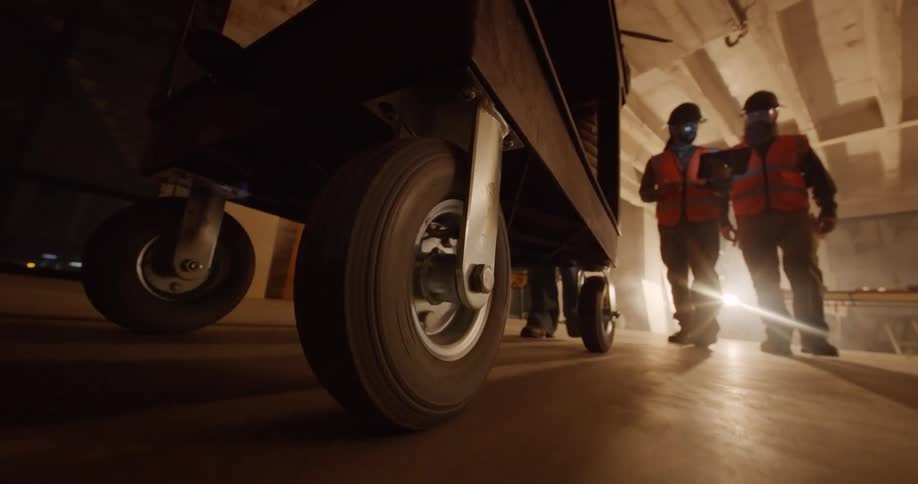 POV from the floor of the wheels of a cart being pushed by a person in a building under construction and being followed by two male workers in hard hats and safety vests.