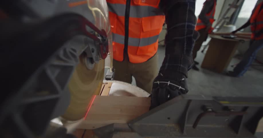 View from a miter saw of a bearded construction worker standing in a building site with other workers around him as he pulls the saw blade down to cut a piece of wood.