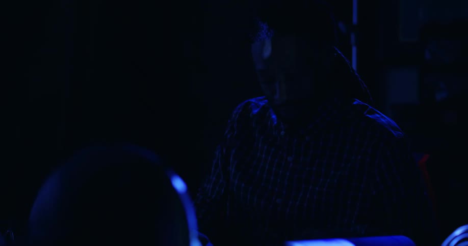 A black man sitting at a desk in a dark room lit only by a blue light projector is speaking and gesturing to someone nearby.