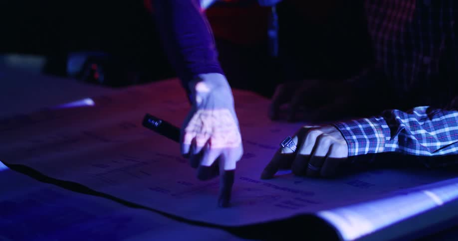 The hands of two different people pointing out details on a project laid out on a table in a dark room lit only a blue-light screen.