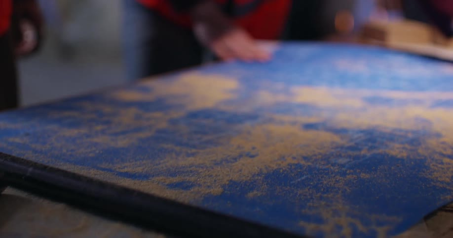View panning over some scattered tools and pieces of wood to a person's hand removing a large piece of paper to reveal a set of blueprints beneath covered in sawdust, which is brushed off by another person in an orange safety vest.