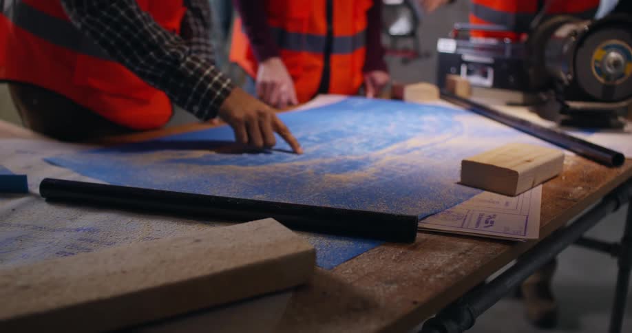 Hands of three engineers pointing to details on the blueprints on the table below them that is covered in sawdust, wood, and tools as they work in an active construction site.