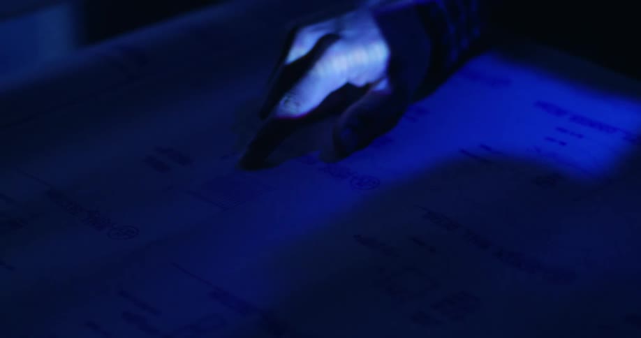 Close up view of a person's hand pointing out words on a piece of paper lit only by a dim blue light reflection from a screen, with a red laser pointer sweeping across at one point.