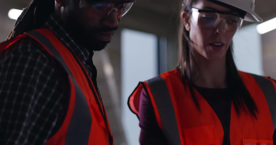 Male and female construction workers wearing hard hats, eye protection, and high-visibility vests focus and collaborate intently on a task in a dimly lit industrial setting