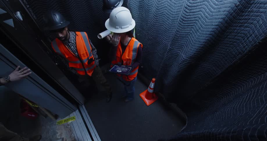 View looking down into a padded, dark elevator as the doors open and 5 construction workers, a woman and 4 men, step in wearing hard hats and safety gear and discussing a building project.