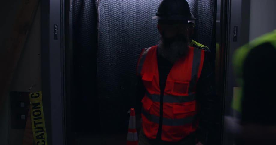 Elevator doors open into a dark room under construction and 5 workers wearing hard hats and safety vest exit the elevator into the room going multiple directions.