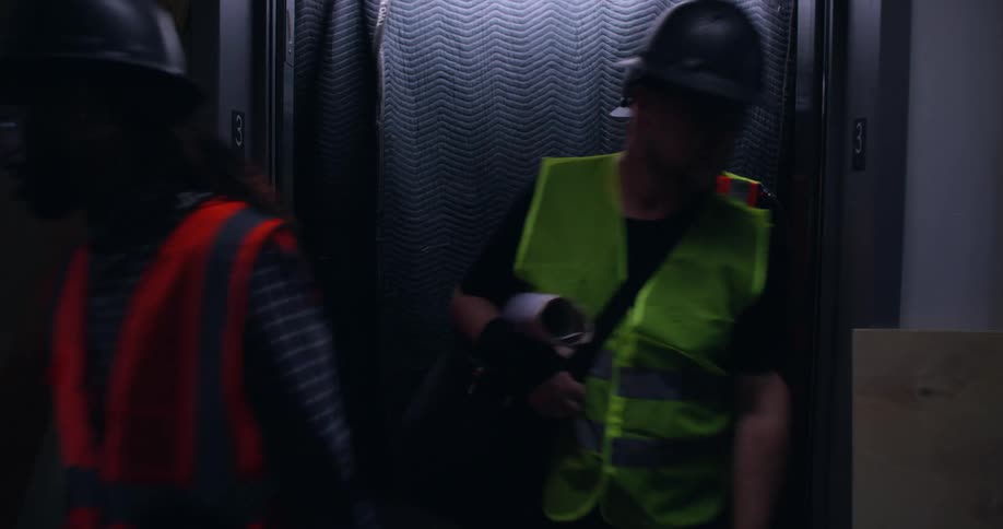 Elevator doors open into a construction area and 5 workers, a woman and 4 men, wearing safety gear and hard hats exit into the dark room.