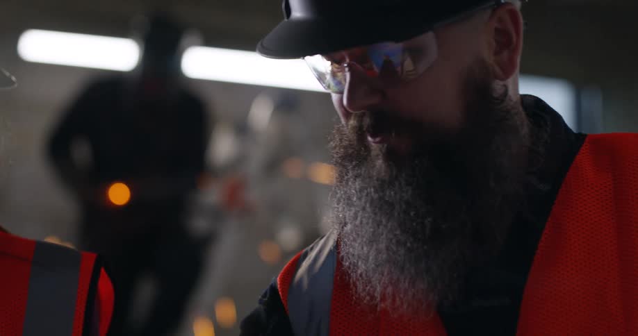 Close up of a bearded, white male construction worker, wearing a hard hat and safety gear, looks down at project plans and discusses with others at an active building site, while a welder sends up sparks in the background.