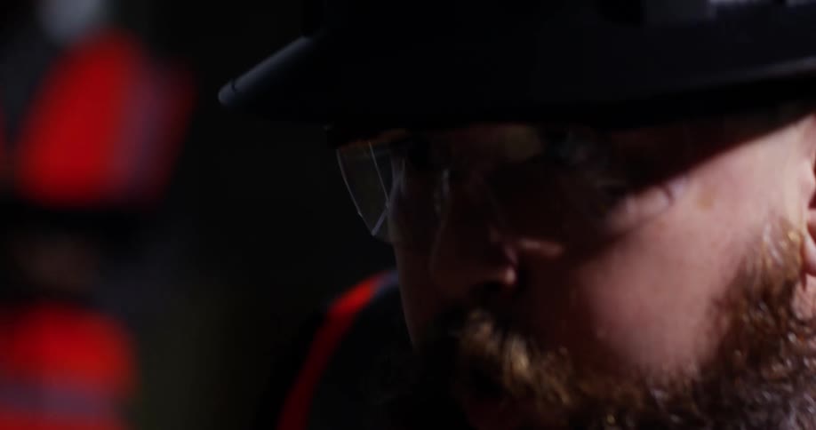 A bearded construction worker leans down to blow the sawdust off of a surface and then walks away as another worker observes in the background.