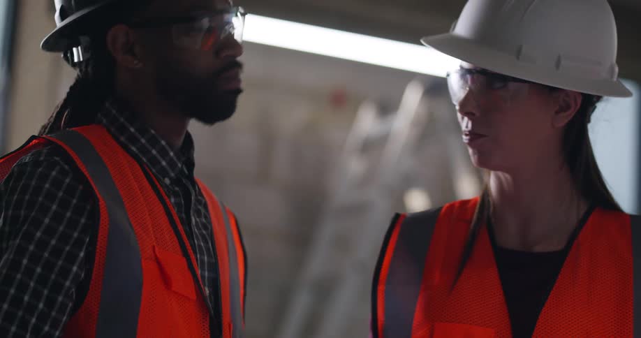 Two construction workers, a black male and white female, wearing safety vests, hard hats, and eye protection, are standing together in an indoor area under construction and discussing building plans.