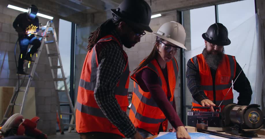 A female civil engineer stands with a foreman and construction supervisor in an indoor construction area discussing project plans laid out in front of them, while a welder works on a ladder in the background giving off sparks.