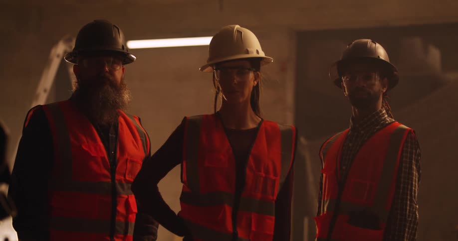Three diverse workers, a woman and two men, standing in the shadows indoors at a construction site while practicing safety first with orange safety vests, eye protection, and hard hats.