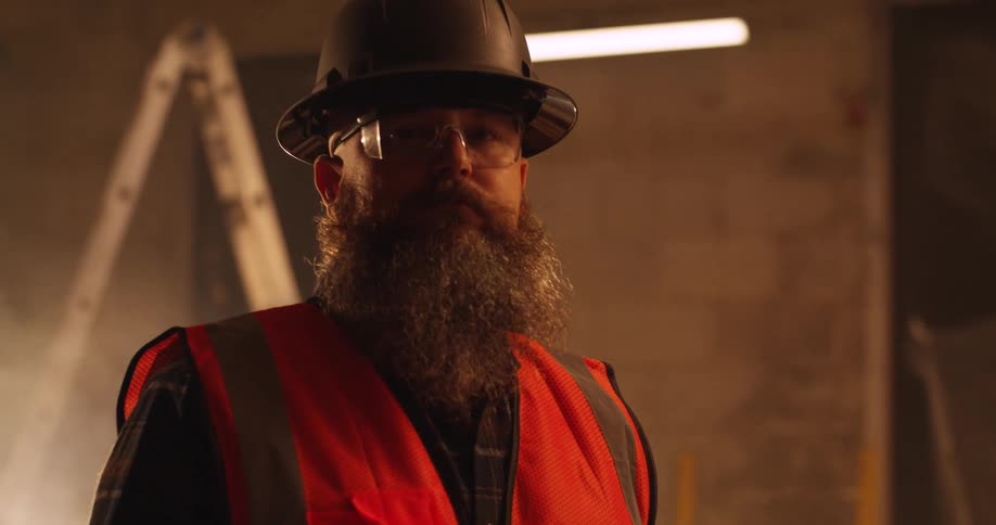 Portrait of a bearded white male contractor standing in a dimly lit and dusty construction zone wearing a hard hat and safety gear.