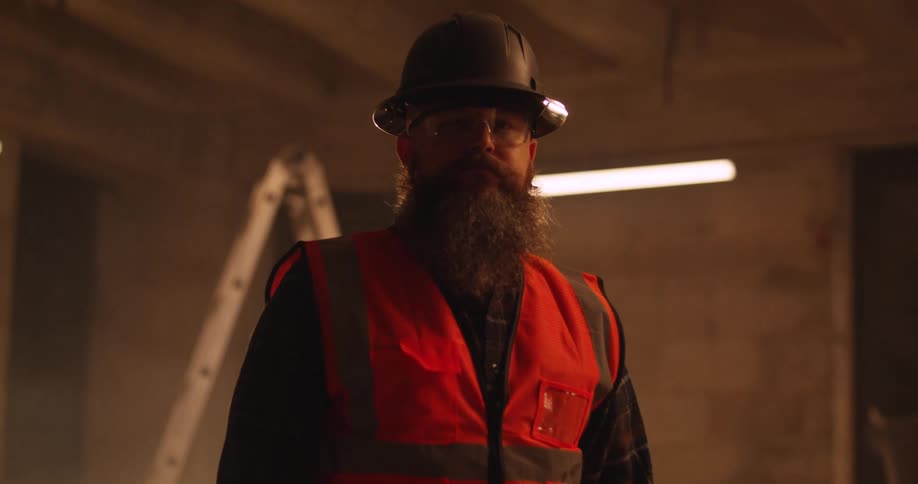 Portrait of a bearded, Caucasian male construction worker wearing a safety vest, eye protection, and a hard hat standing in a dusty, dimly lit, indoor construction zone.