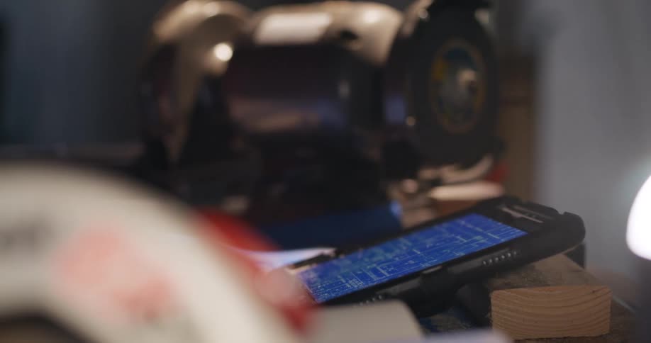 View from behind a blurred object of a tablet leaning on a raw piece of wood with technical building project blueprints displayed on the screen while the hands of two people interact.