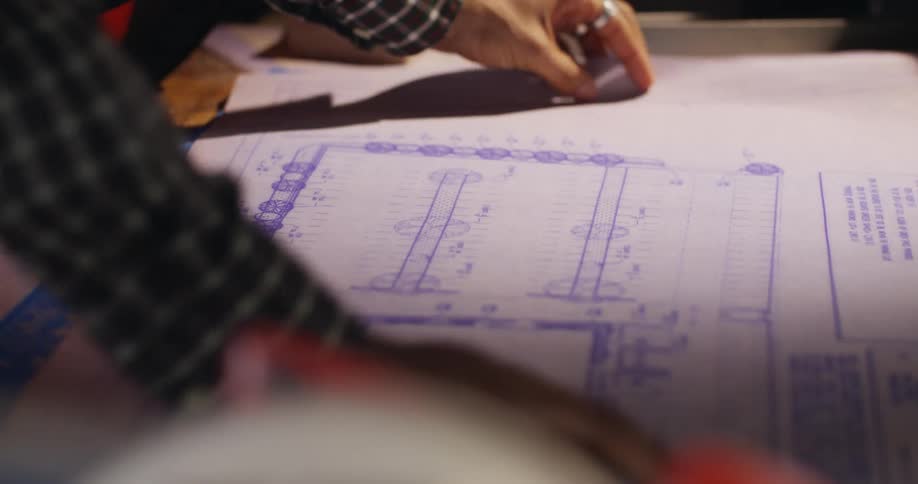 A pair of hands unrolls some blueprints and then points to technical details on the printed architectural design.