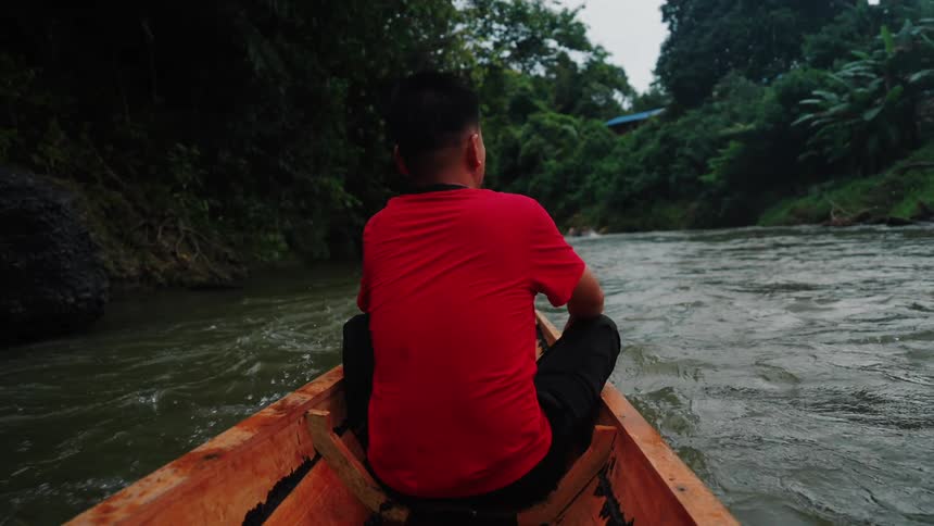 Man in a red shirt sitting on the bow of a wooden canoe sailing down a river surrounded by dense vegetation
