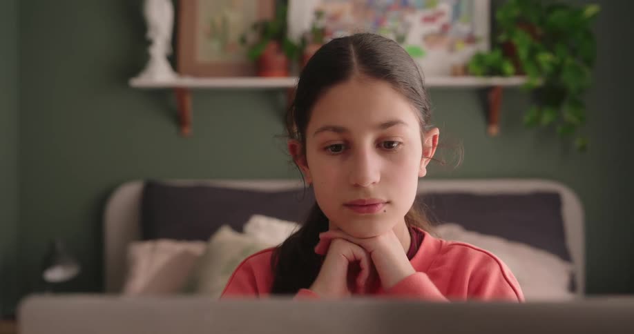 A girl wearing an orange sweatshirt sits in front of her bed using a computer in her room