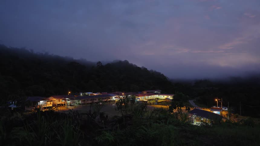 A cluster of dimly lit buildings nestles in a dark valley surrounded by misty hills under a twilight sky