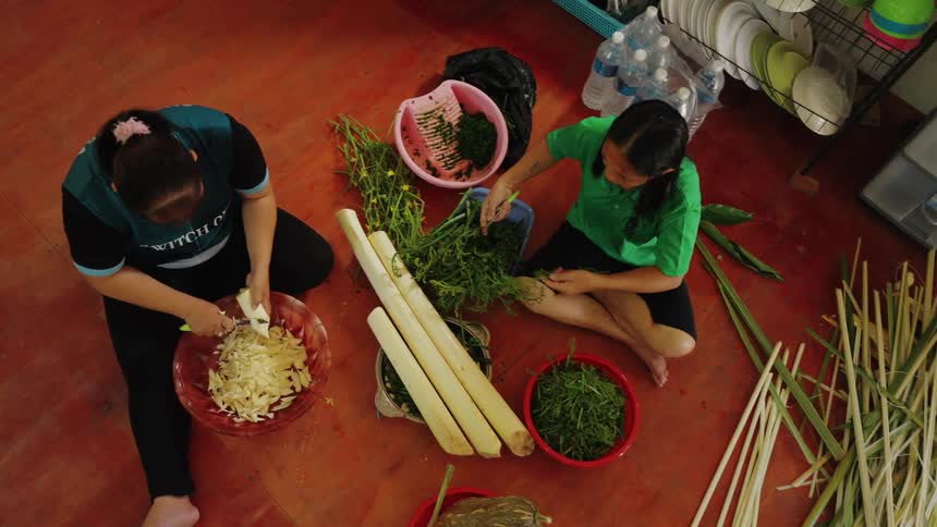 Two individuals are seated on the floor preparing food with one person peeling bamboo shoots into a bowl and the other person sorting leafy greens surrounded by various bowls bamboo shoots and bottled water