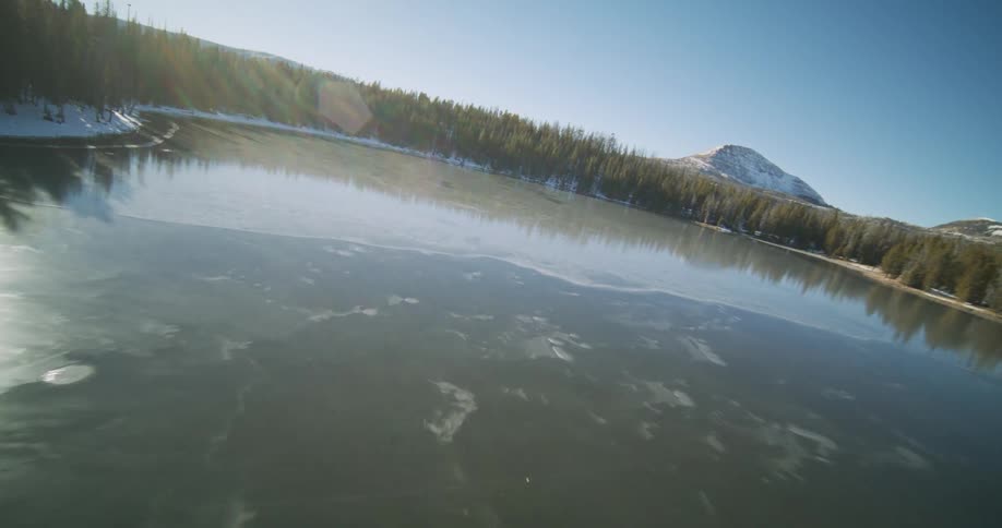 A partially frozen lake surrounded by evergreen forests and distant mountains under a clear blue sky