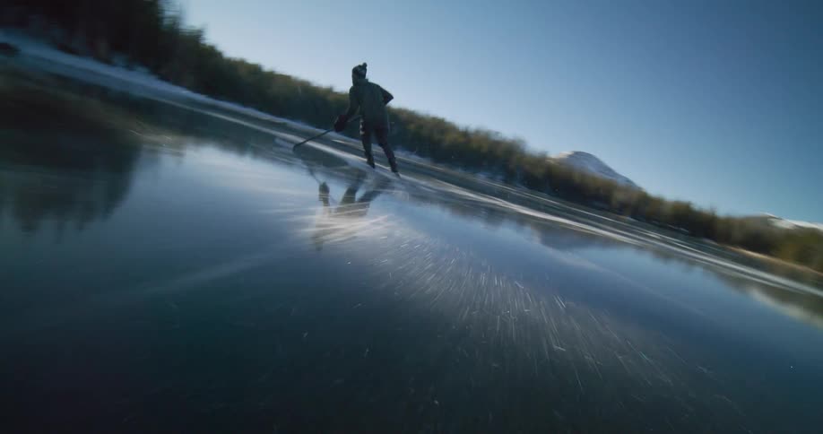 A person ice skates with a hockey stick on a frozen lake surrounded by snowy trees and mountains under a sunny sky