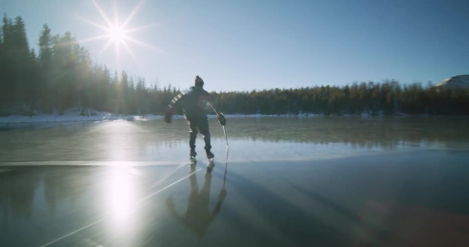 An aerial shot captures a figure ice skating on a partly frozen lake surrounded by snow dusted evergreens under a brilliant sun