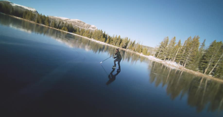 Hockey athlete practices skating across a reflective pond surrounded by a conifer forest on a sunny day