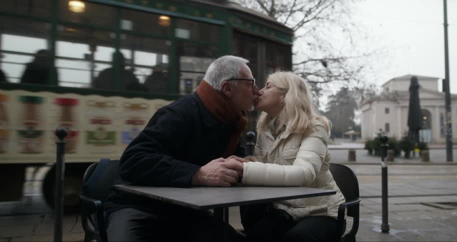 Elderly couple sitting together holding hands at an outdoor table with a cable car passing by in the background