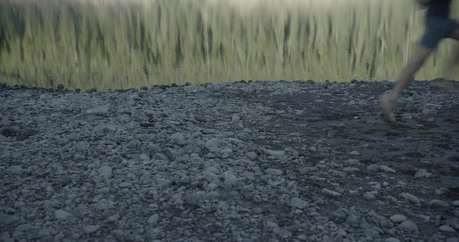 A person is running on a rocky shore with a calm lake and a reflection of tall grass or reeds in the background