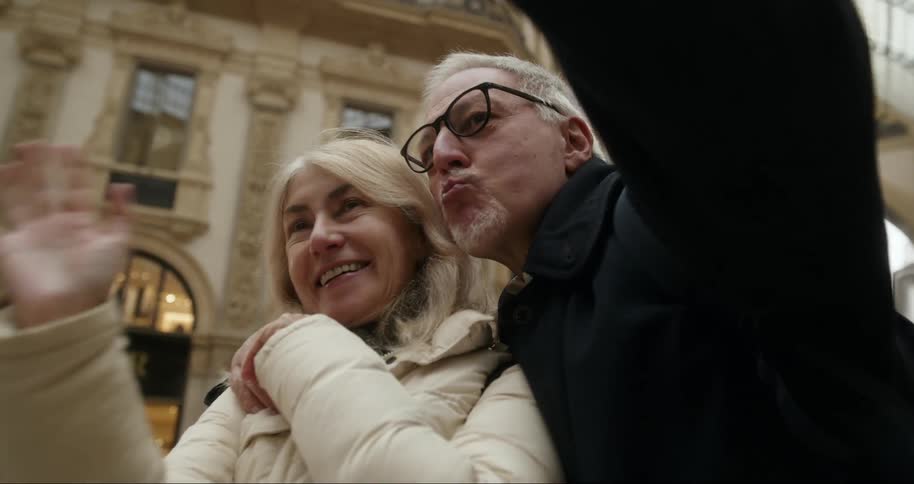 Inside a grand building a couple is joyfully posing for a photo as the husband playfully blows kisses while holding a smartphone to capture the moment and his wife waves and laughs
