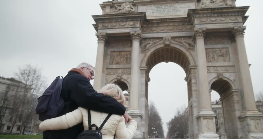 Older couple hugs each other while walking towards a historic arc de triomphe du carrousel on a winter day